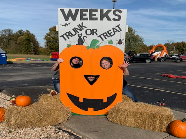 Painted jack-o-lantern with cutouts at the eyes and nose for photos. It reads 'Weeks Trunk or Treat' above the pumpkin.